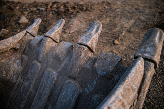 Worn excavator bucket teeth and adapters on a dirt job site — aftermarket ground engaging tools, bucket teeth, adapters, and side cutters for construction and mining equipment