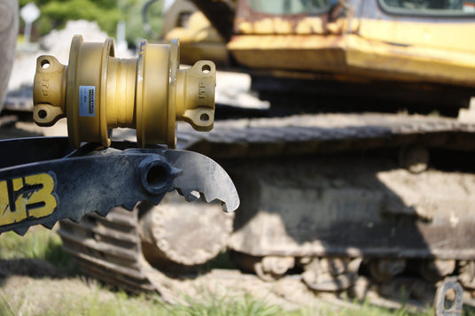ITP-branded track roller and bucket tine in front of a heavy excavator — aftermarket rollers, hardware, wear parts, and additional components for construction equipment